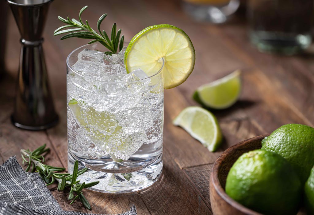 Gin and tonic cocktail with lime slices, rosemary and ice on a rustic wooden table